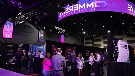 A large room with lots of booths set up, each from a different technology company. People in business-casual clothing are walking around and socializing.