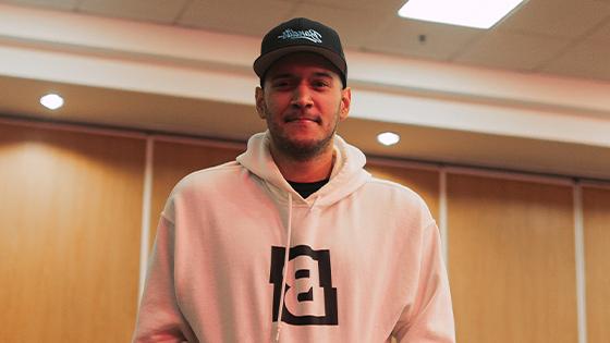 A man standing against a wood paneled wall in a white hooded sweatshirt featuring the Bandits logo and a black Bandits Gaming snapback cap.